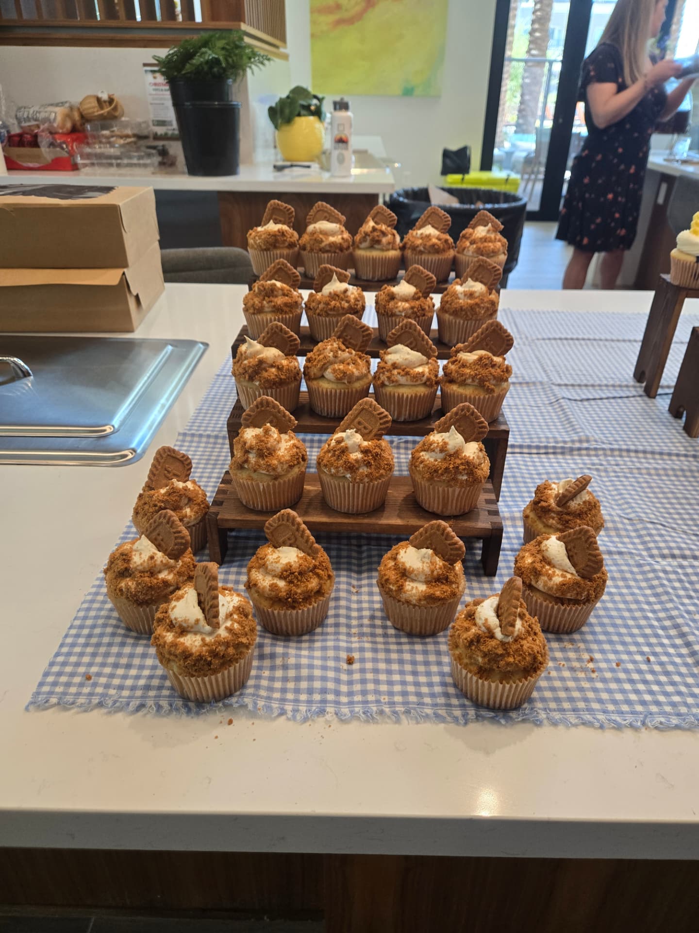 Tiered wooden stand holding rows of Biscoff crumb cupcakes on a blue gingham runner